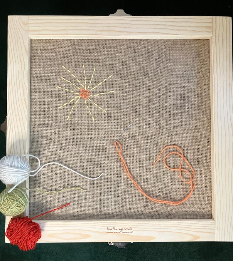 May include: A framed embroidery project featuring a sun design stitched with yellow and orange thread on a tan fabric. Several balls of yarn in white, green, and red are in the lower left corner. The frame is made of light-colored wood.