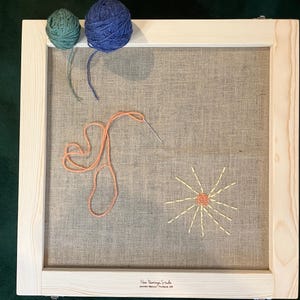 May include: A wooden embroidery frame with a burlap fabric stretched inside. The frame holds an unfinished embroidery project with a sun design and a heart shape. Two balls of yarn, one teal and one blue, sit at the top. The text "Flower Paintings Studio" is at the bottom.