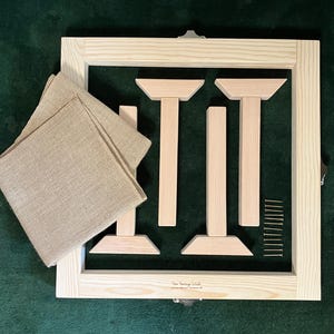 May include: A wooden embroidery frame kit with a square frame, wooden supports, screws, and tan fabric. The frame is made of light-colored wood and includes metal clasps. The fabric is folded and placed on the frame.