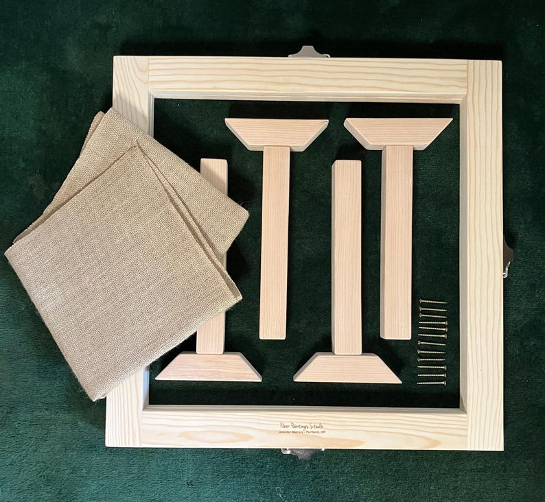 May include: A wooden embroidery frame kit with a square frame, wooden supports, screws, and tan fabric. The frame is made of light-colored wood and includes metal clasps. The fabric is folded and placed on the frame.