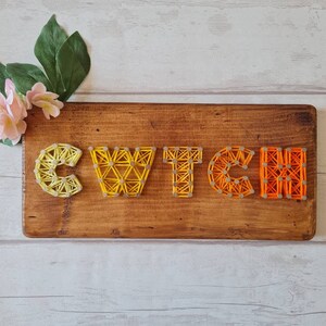 May include: A wooden sign with the word "WITCH" spelled out in string art. The string is yellow, orange, and red, creating a gradient effect.