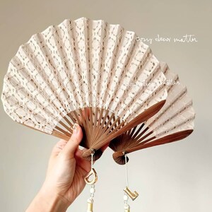 May include: Two folding fans with cream-colored lace fabric and dark brown wooden ribs. Each fan features a hanging charm with a gold-tone letter, a small white bead, and a long, light beige tassel. The text "cozy decor matter" is visible.