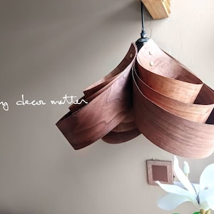 May include: A wooden pendant light fitting with a unique, layered design. The light is made of thin strips of wood that are curved and layered to create a flowing, organic shape. The light is hanging from a ceiling hook and is illuminated by a light bulb.