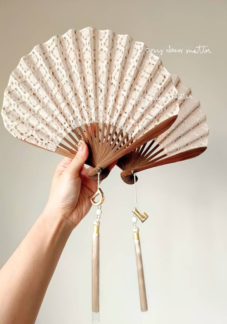 May include: Two folding fans with cream-colored lace fabric and dark brown wooden ribs. Each fan features a hanging charm with a gold-tone letter, a small white bead, and a long, light beige tassel. The text "cozy decor matter" is visible.