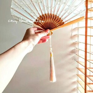 May include: A folding hand fan featuring a light blue fabric canopy with a white lace overlay and a wooden frame. The fan includes a long, light-colored decorative tassel hanging from the base. The text "cozy dew atelier" is visible on the fan.