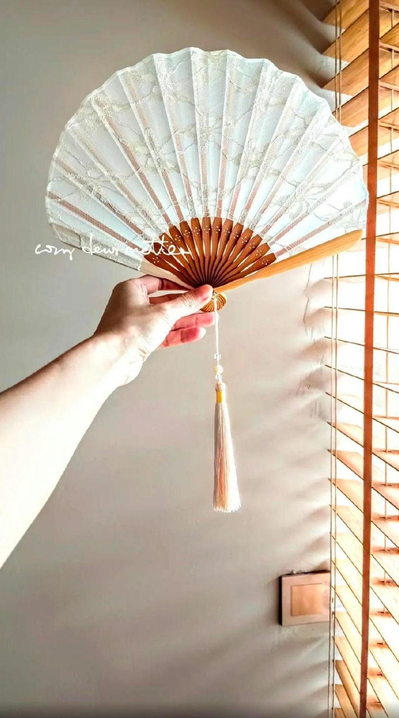 May include: A folding hand fan featuring a light blue fabric canopy with a white lace overlay and a wooden frame. The fan includes a long, light-colored decorative tassel hanging from the base. The text "cozy dew atelier" is visible on the fan.