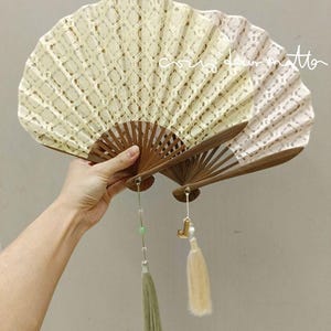 May include: Two folding hand fans with brown wooden ribs and cream-colored lace fabric featuring a diamond pattern. One fan has a green tassel with a small bead, and the other has a cream tassel with a gold-toned letter charm attached to the base.