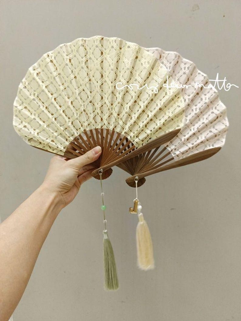 May include: Two folding hand fans with brown wooden ribs and cream-colored lace fabric featuring a diamond pattern. One fan has a green tassel with a small bead, and the other has a cream tassel with a gold-toned letter charm attached to the base.