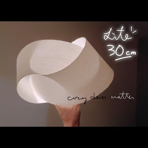 May include: A white, paper-like lampshade with a unique, folded design. The lampshade is 30 inches in diameter and has a text overlay that reads "Lite' 30cm cozy decor matter".