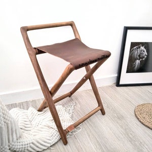 May include: A folding wooden stool with a brown leather seat. The frame is made of dark wood, and the seat is a rich brown color. The stool is designed to fold flat for easy storage. The words "cozy dear mother" are written on the seat.