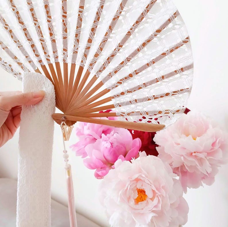 May include: A handheld folding fan featuring light brown wooden ribs and a white lace canopy. A gold-toned letter V charm hangs from the base of the fan, attached to a thin white ribbon and a light pink tassel. The fan is open in a semi-circle.