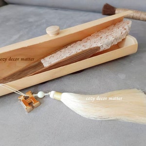 May include: A folded beige lace hand fan with wooden ribs rests inside a light-colored wooden box. A gold-toned letter H charm, a small white bead, and a long cream-colored silk tassel are attached to the fan. The text cozy decor matter is visible.