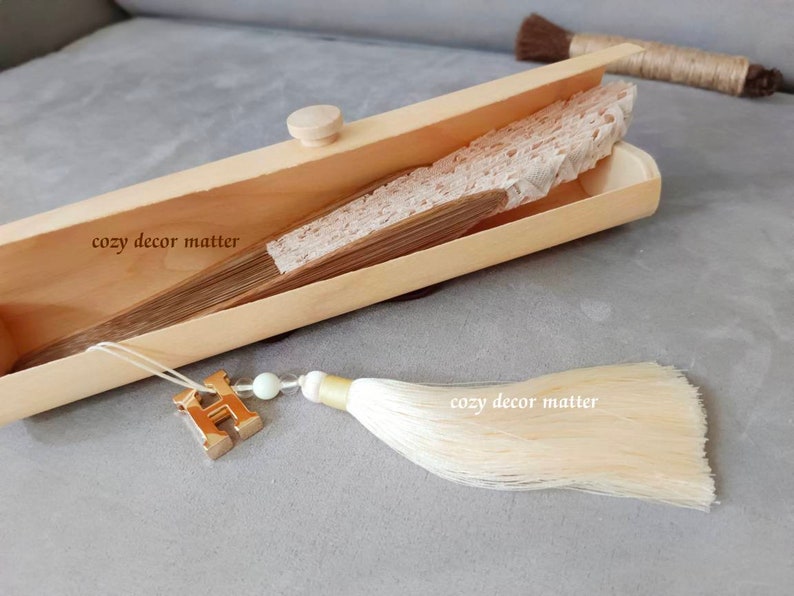 May include: A folded beige lace hand fan with wooden ribs rests inside a light-colored wooden box. A gold-toned letter H charm, a small white bead, and a long cream-colored silk tassel are attached to the fan. The text cozy decor matter is visible.