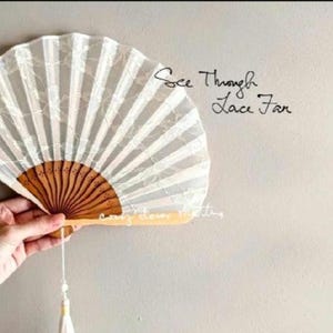 May include: A white lace folding fan with a brown wooden frame and a hanging tassel. The fan features a semi-transparent floral pattern and the text See Through Lace Fan printed in black script on the right side of the fan surface.