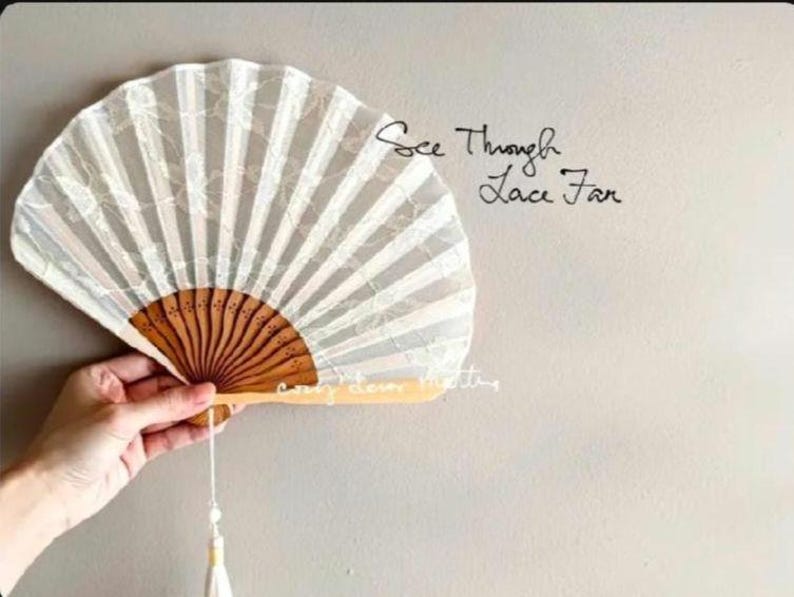 May include: A white lace folding fan with a brown wooden frame and a hanging tassel. The fan features a semi-transparent floral pattern and the text See Through Lace Fan printed in black script on the right side of the fan surface.