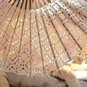 May include: A folding hand fan featuring a cream-colored lace fabric with an intricate geometric pattern and dark wooden ribs. The fan is partially open, displaying the delicate lace texture and the text "cozy deer mater" written in white script.