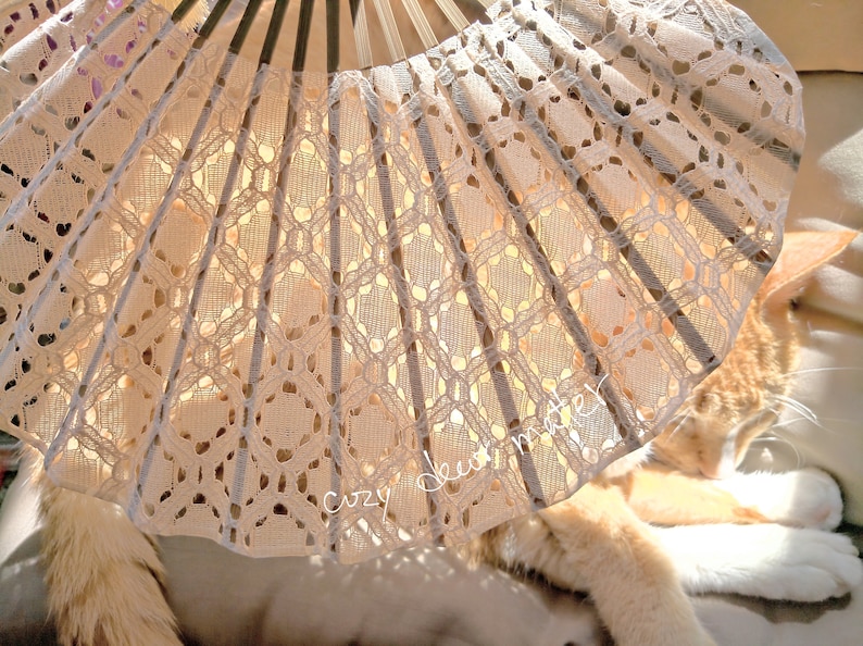 May include: A folding hand fan featuring a cream-colored lace fabric with an intricate geometric pattern and dark wooden ribs. The fan is partially open, displaying the delicate lace texture and the text "cozy deer mater" written in white script.