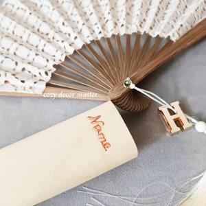 May include: A folding hand fan with white lace fabric and brown wooden ribs. A light-colored wooden case features the word Name in copper-colored script. A gold-toned letter H charm and a cream-colored tassel are attached to the fan handle.