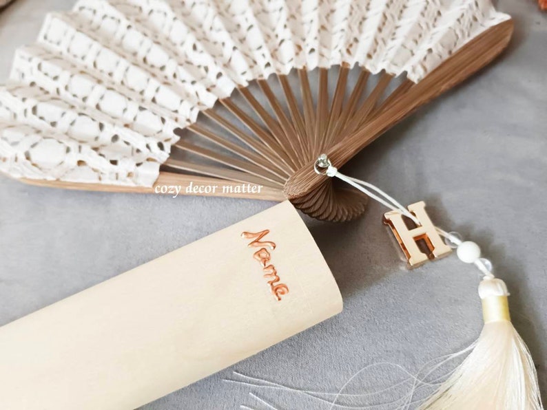 May include: A folding hand fan with white lace fabric and brown wooden ribs. A light-colored wooden case features the word Name in copper-colored script. A gold-toned letter H charm and a cream-colored tassel are attached to the fan handle.