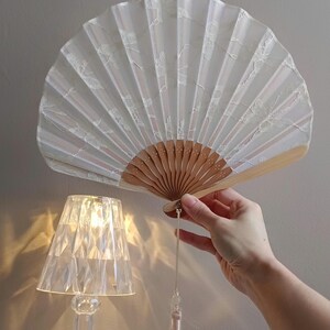 May include: A folding hand fan featuring a white fabric canopy with a delicate white lace overlay pattern. The fan is constructed with light brown wooden ribs and includes a long, light-colored decorative tassel hanging from the base handle.