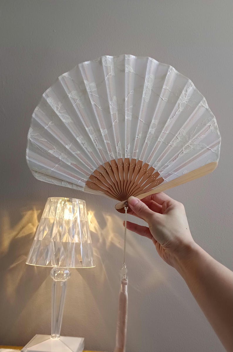 May include: A folding hand fan featuring a white fabric canopy with a delicate white lace overlay pattern. The fan is constructed with light brown wooden ribs and includes a long, light-colored decorative tassel hanging from the base handle.