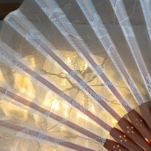 May include: A handheld folding fan featuring a cream-colored lace fabric overlay with a delicate floral pattern. The fan is constructed with light brown wooden ribs that radiate from a central pivot point, each rib displaying small cut-out details.