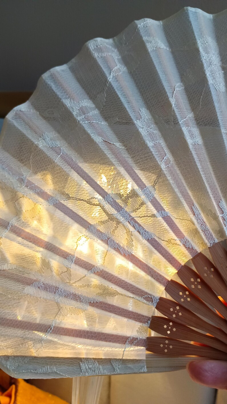 May include: A handheld folding fan featuring a cream-colored lace fabric overlay with a delicate floral pattern. The fan is constructed with light brown wooden ribs that radiate from a central pivot point, each rib displaying small cut-out details.
