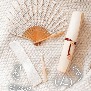May include: A white lace folding fan with a wooden frame and a hanging tassel, a white lace sleeve, and a rolled light-colored paper scroll tied with a dark red ribbon featuring a small gold-toned button and a hanging letter S charm.