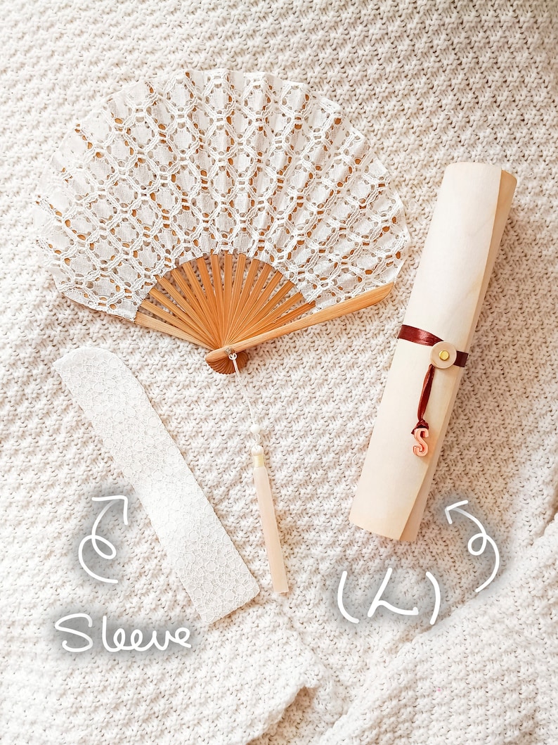 May include: A white lace folding fan with a wooden frame and a hanging tassel, a white lace sleeve, and a rolled light-colored paper scroll tied with a dark red ribbon featuring a small gold-toned button and a hanging letter S charm.