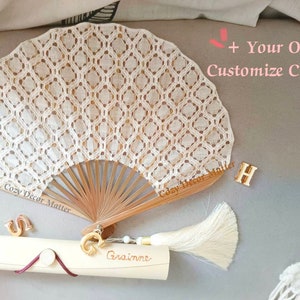 May include: A beige lace hand fan with a wooden frame and a cream-colored tassel. The fan features a diamond pattern. Included are gold-toned letter charms S, G, and H, and a cylindrical tube with the name Grainne and text + Your Own Customize Charm.
