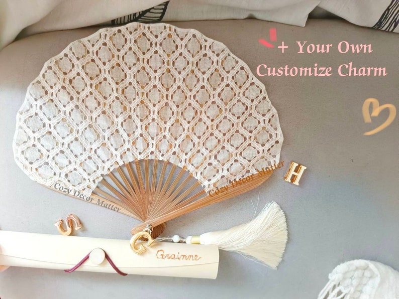 May include: A beige lace hand fan with a wooden frame and a cream-colored tassel. The fan features a diamond pattern. Included are gold-toned letter charms S, G, and H, and a cylindrical tube with the name Grainne and text + Your Own Customize Charm.