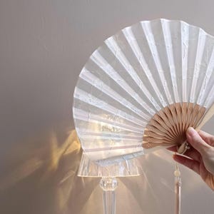 May include: A white folding hand fan with a translucent, textured fabric leaf and light-colored wooden ribs. The fan features a delicate, repeating pattern on the fabric and a matching light-colored tassel hanging from the base of the handle.