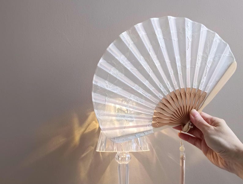 May include: A white folding hand fan with a translucent, textured fabric leaf and light-colored wooden ribs. The fan features a delicate, repeating pattern on the fabric and a matching light-colored tassel hanging from the base of the handle.
