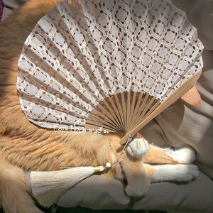 May include: A handheld folding fan featuring a beige lace canopy with a geometric pattern, light brown wooden ribs, and a long beige tassel with a small white bead. The text "Cozy deco matter" is printed in white on the fan's wooden ribs.