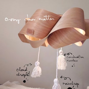 May include: A wooden light fitting with a unique, flowing design. The fitting is suspended from the ceiling with white tassels hanging from it. The text "Cozy, dear matter" is written on the wall behind the fitting.