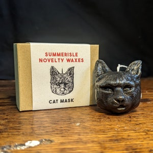 May include: A black wax candle shaped like a cat's head. The candle is packaged in a box with the text "Summerisle Novelty Waxes" and "Cat Mask" printed on it.