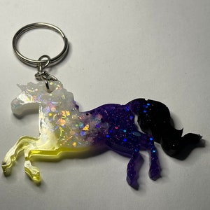 May include: A white, yellow, purple, and black unicorn keychain with glitter embedded in the resin. The unicorn is standing on its hind legs with its front legs raised.