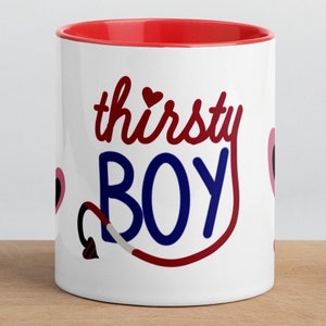 Puede incluir: Taza de cerámica blanca con borde rojo. La taza tiene un diseño rojo y azul con el texto "thirsty BOY" y un corazón rojo.
