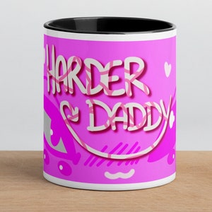 May include: A pink ceramic mug with a black rim. The mug has a pink and white graphic that says "Harder Daddy" with hearts and other designs.