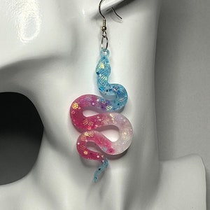 Peut inclure: Une paire de boucles d'oreilles pendantes avec un motif de serpent bleu et rose dégradé avec des accents de paillettes. Le serpent est enroulé dans une pose réaliste et les boucles d'oreilles ont des crochets argentés.