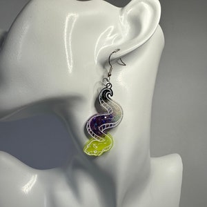 May include: A pair of dangle earrings featuring a black, purple, and yellow swirl design. The swirl is filled with iridescent glitter. The earrings are made of resin and have silver hooks.