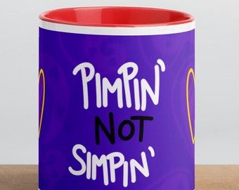 May include: A white ceramic mug with a red interior and a purple exterior. The mug features the text "Pimpin' Not Simpin'" in white, with yellow heart-shaped designs on either side. The mug sits on a wooden surface.