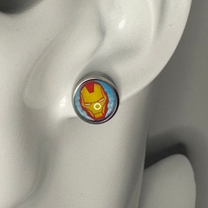 Puede incluir: Un par de pendientes de botón de plata con una imagen redonda de la cara de Iron Man. La imagen es amarilla, roja y azul.
