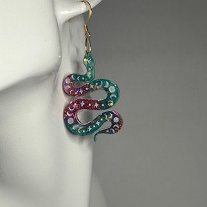 Op de afbeelding: Een paar hangende oorbellen met een groene en roze slang met witte maan- en steraccenten. De slang is gemaakt van hars en heeft een glinsterende afwerking.