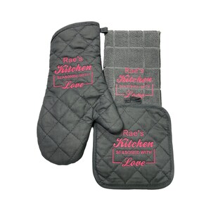 May include: Black quilted oven mitt and pot holder set with a gray kitchen towel. All three items have the text "Rae's Kitchen Seasoned with Love" embroidered in pink.