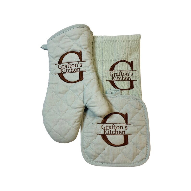 May include: A set of three kitchen accessories in a light green color with a quilted texture. The set includes a pot holder, an oven mitt, and a tea towel. All three items feature the embroidered text "Grafton's Kitchen" with a large "G" monogram.