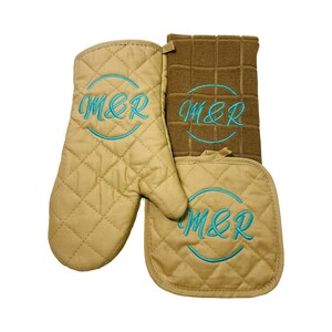 May include: A tan quilted oven mitt, pot holder, and dish towel set. Each piece is embroidered with the letters "M&R" in a teal script within a teal circle. The dish towel is a darker brown color.