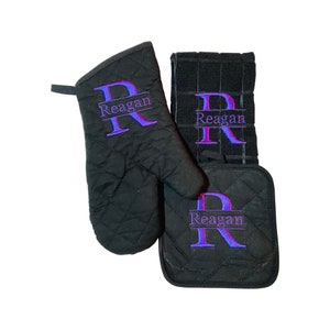 May include: Black quilted oven mitt, pot holder, and kitchen towel set with the name "Reagan" embroidered in purple on each piece.