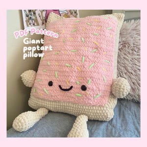 May include: A rectangular crochet pillow shaped like a pastry with a pink frosted top featuring scattered green and yellow sprinkles. The pillow has a cream-colored crust, two small arms, two legs, and a smiling face with black eyes and a mouth.