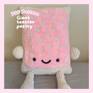 May include: A rectangular crochet plush toy shaped like a toaster pastry with a cream-coloured base and a pink frosted top featuring green and yellow sprinkles. The face has black safety eyes and a stitched smile. Text reads: PDF Pattern Giant toaster pastry.